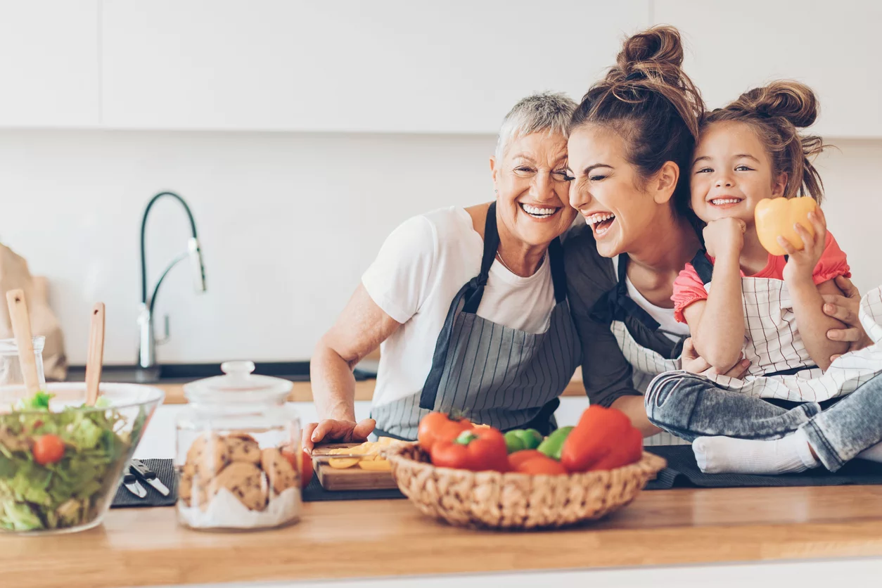 Drei Generationen – Großmutter, Mutter und Kind – stehen lachend an einer massiven Küchenarbeitsplatte aus Eichenholz.